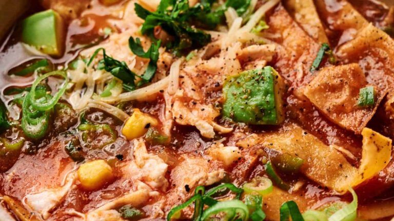 Close-up of a bowl of chicken tortilla soup with avocado, tortilla strips, corn, shredded cheese, green onions, and cilantro in a tomato-based broth.