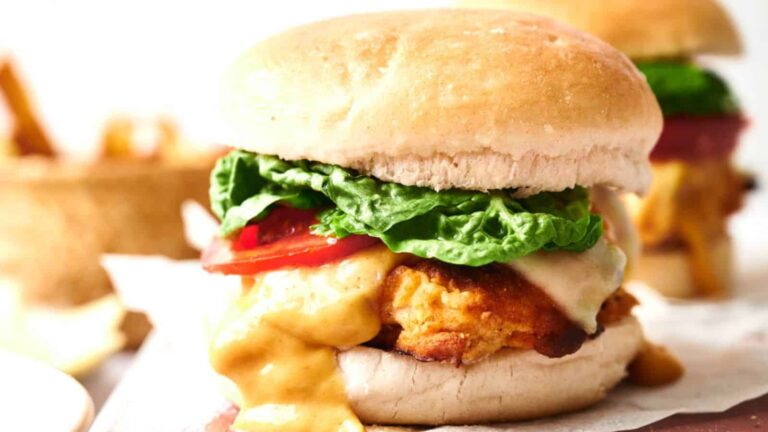 A fried chicken sandwich with melted cheese, lettuce, and tomato on a bun, with sauce dripping down the side.