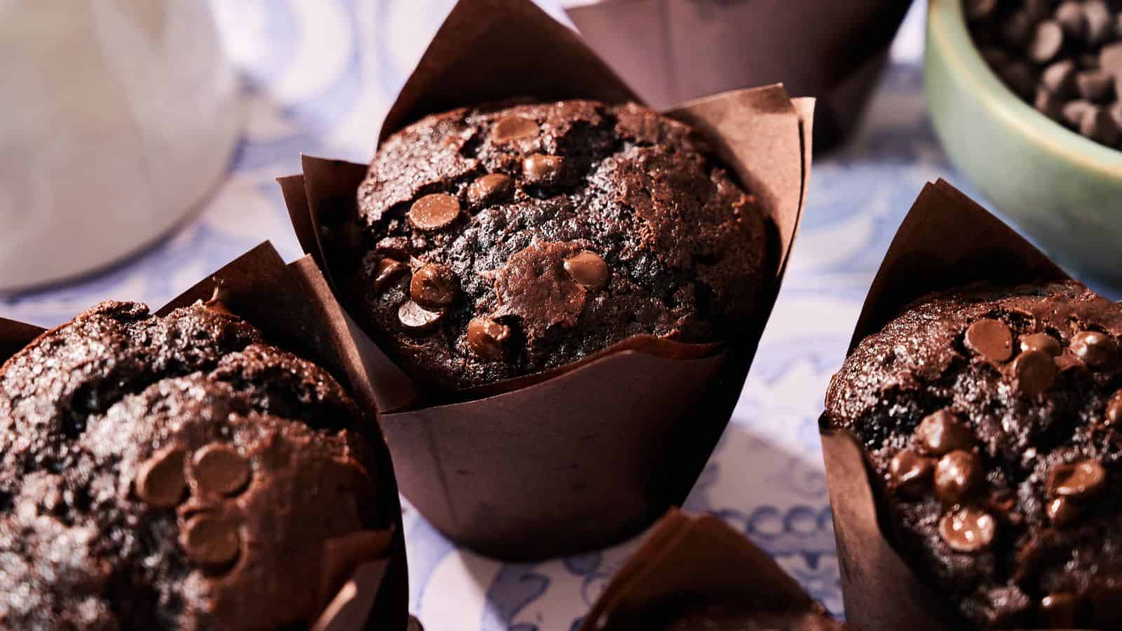 Close-up of three chocolate muffins in brown paper wrappers, topped with chocolate chips, with part of a bowl of chocolate chips visible in the background.