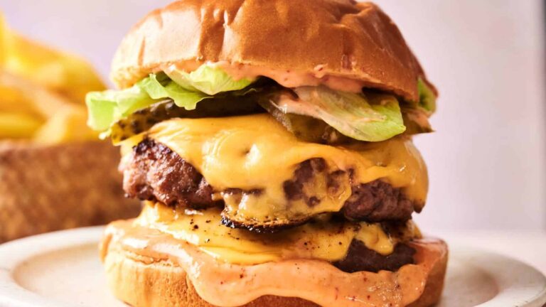 A double cheeseburger with lettuce, pickles, cheese, and sauce in a toasted bun, served on a white plate with fries in the background.
