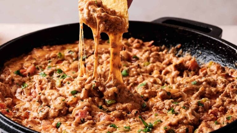 A skillet filled with cheesy ground beef dip, garnished with herbs, as a tortilla chip is dipped in, stretching melted cheese.