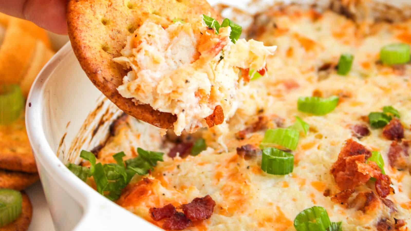 Cracker with creamy dip featuring bacon bits and green onions being lifted from a dish.