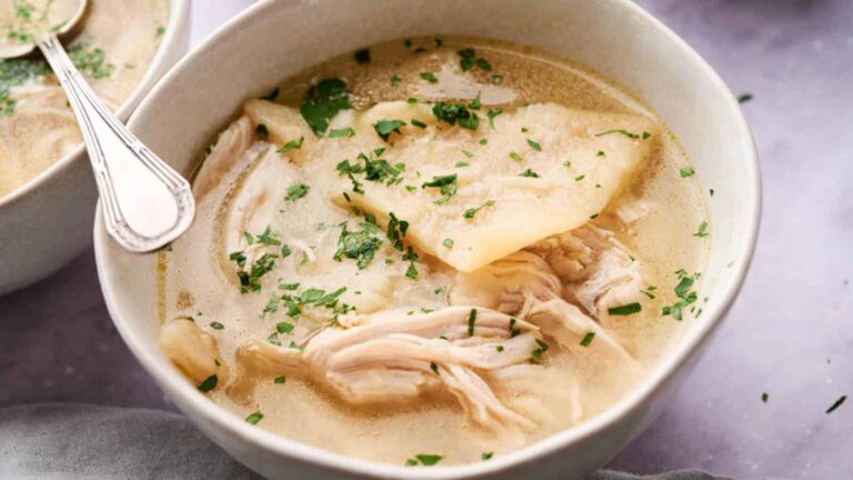A bowl of chicken and dumplings soup with shredded chicken, dumplings, and chopped herbs, served with a spoon.