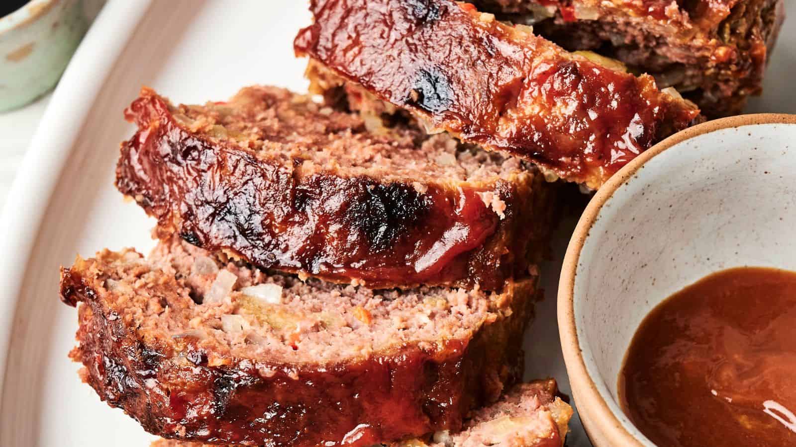 Three slices of glazed meatloaf are arranged on a plate next to a bowl of brown sauce.