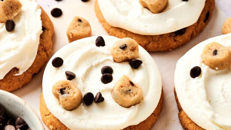 Frosted cookies topped with small chunks of cookie dough and chocolate chips, arranged on a white surface.