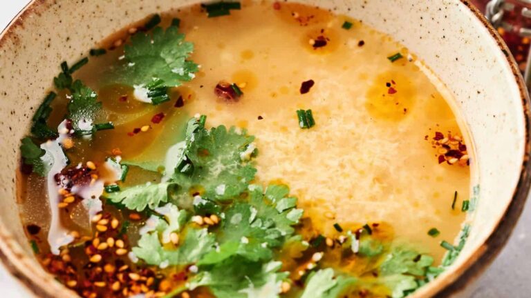 A close-up of a bowl of egg drop soup garnished with cilantro, chopped chives, sesame seeds, chili flakes, and a touch of oil.