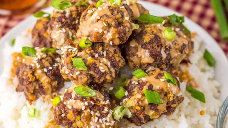 A plate of rice topped with several beef meatballs, garnished with sesame seeds, chopped green onions, and a creamy sauce.