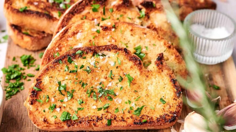 Several slices of toasted garlic bread topped with chopped parsley and coarse salt are arranged on a wooden board. A small bowl of salt and garlic cloves are visible in the background.