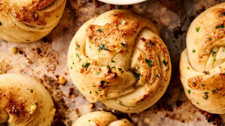 Close-up of baked garlic knots topped with herbs and melted butter on a parchment-lined baking sheet.