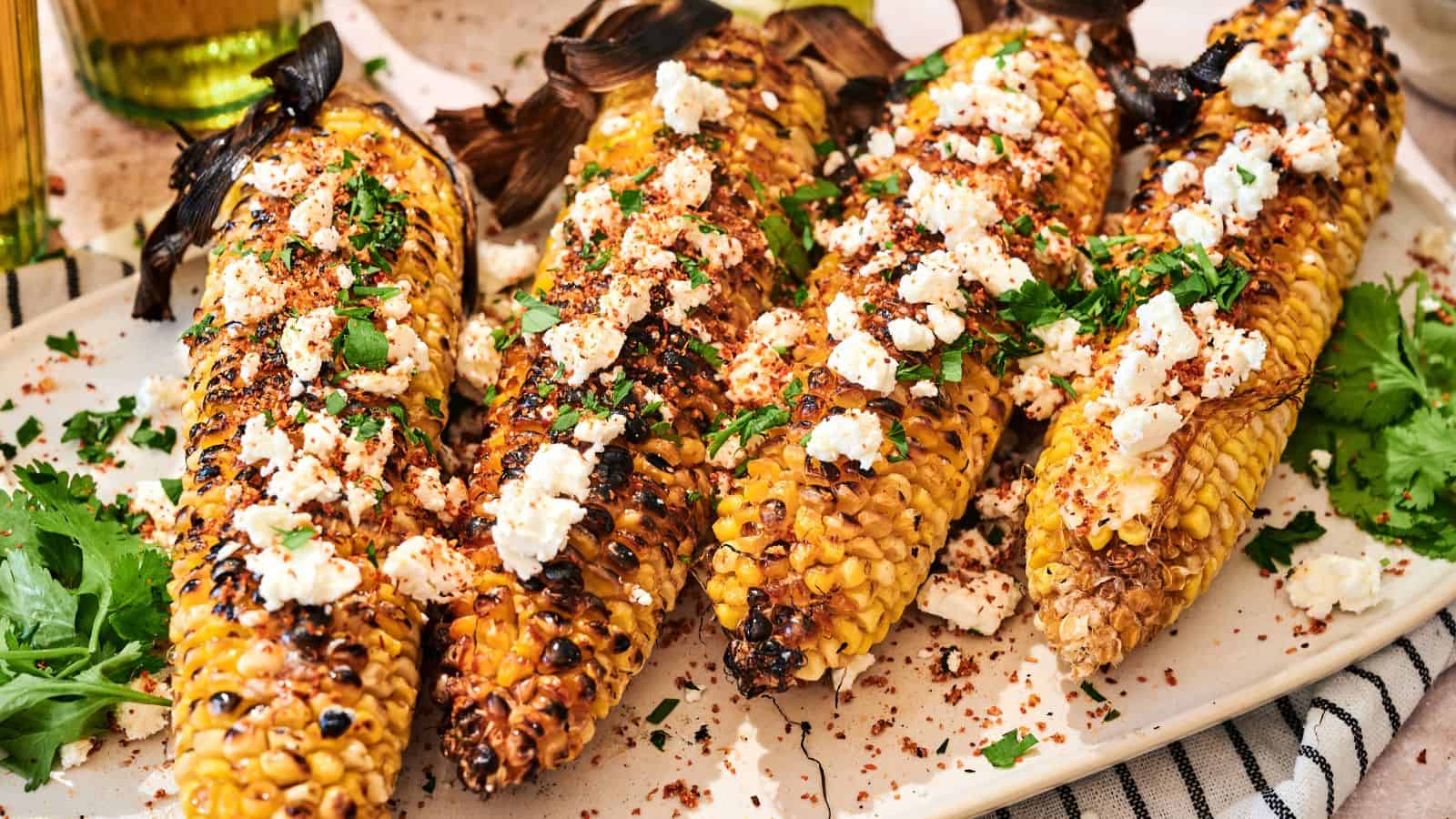 Four grilled corn cobs topped with crumbled cheese, chopped herbs, and spices are arranged on a white plate.
