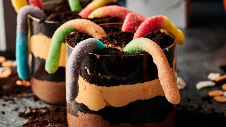 Two layered dessert cups with chocolate pudding, crushed cookies, creamy filling, and gummy worms on top.