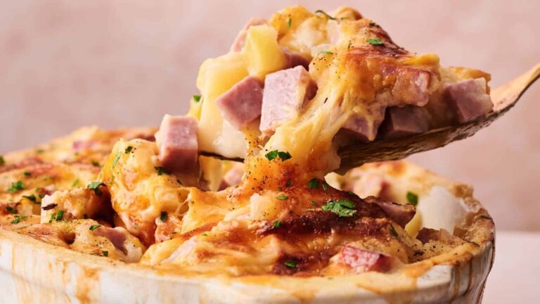 A close-up of a cheesy casserole with diced ham and potatoes being served with a wooden spoon.