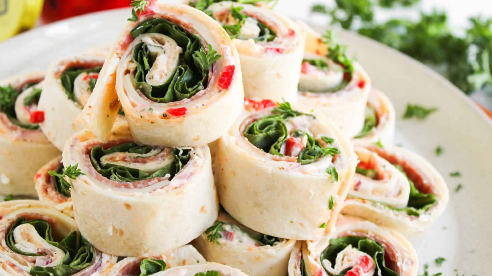 A plate of tortilla pinwheel sandwiches filled with spinach, cream cheese, red peppers, and herbs, neatly stacked and garnished with parsley.