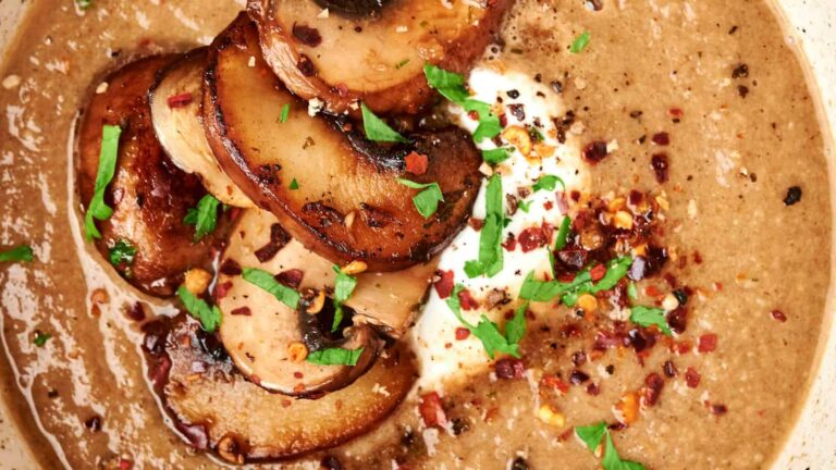 A bowl of creamy mushroom soup topped with sautéed mushroom slices, chopped parsley, red pepper flakes, and a dollop of cream.