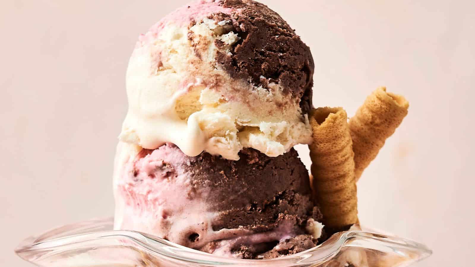 Two scoops of Neapolitan ice cream in a glass dish, featuring chocolate, vanilla, and strawberry flavors, served with two rolled wafer cookies.