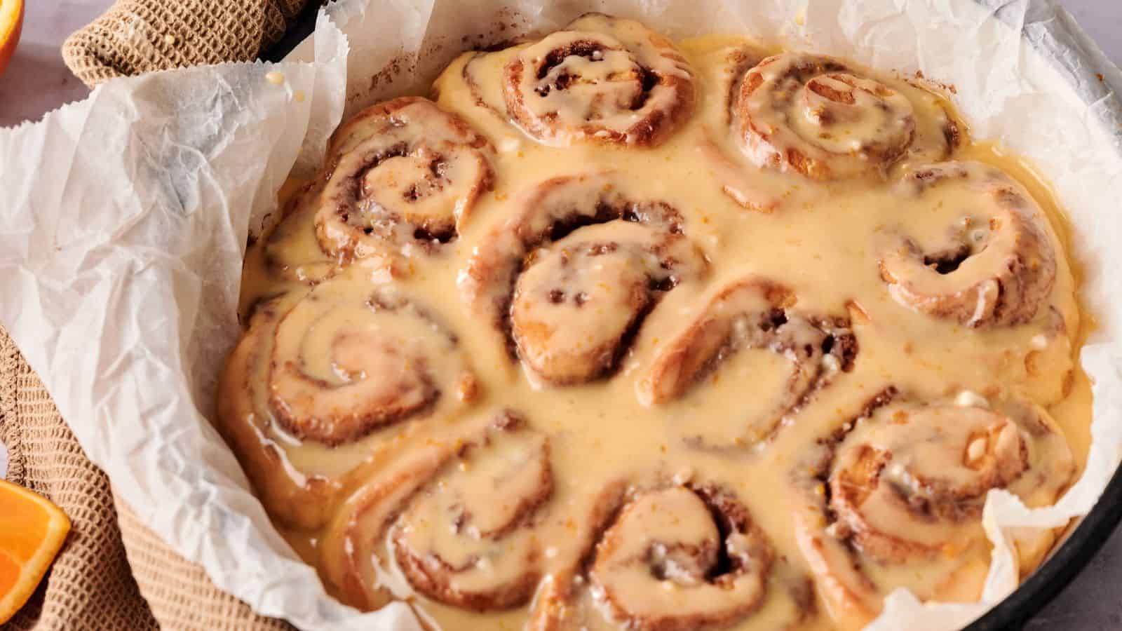 A round baking dish lined with parchment paper holds several cinnamon rolls covered in a thick layer of icing.