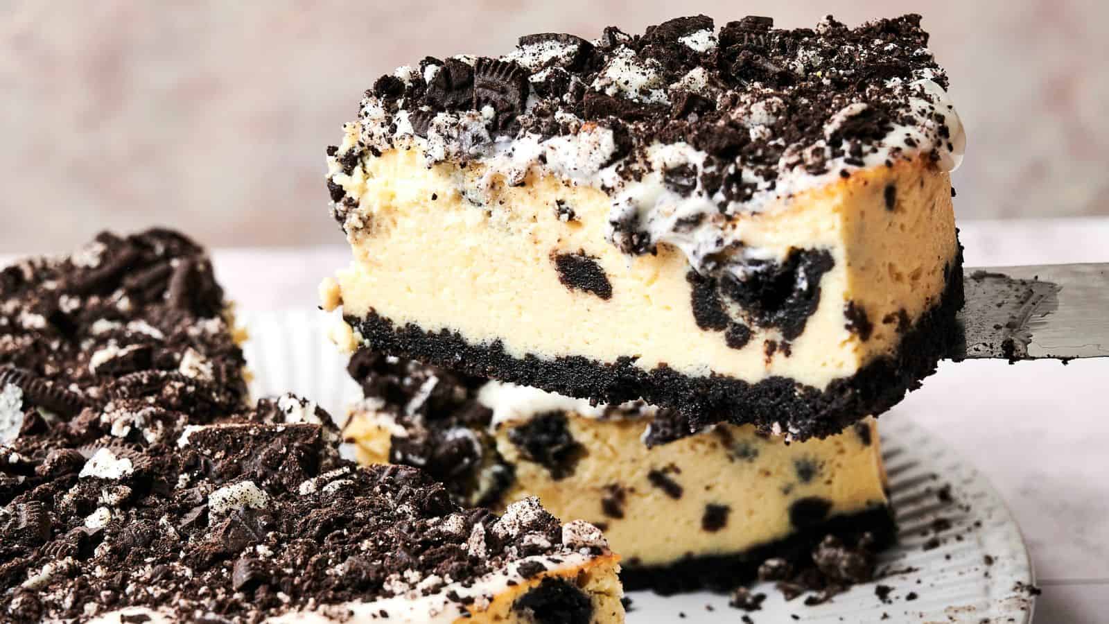 A slice of cookies and cream cheesecake is being lifted from a plate. The cake has a crumbly cookie crust, creamy filling with cookie pieces, and a topping of crushed cookies.