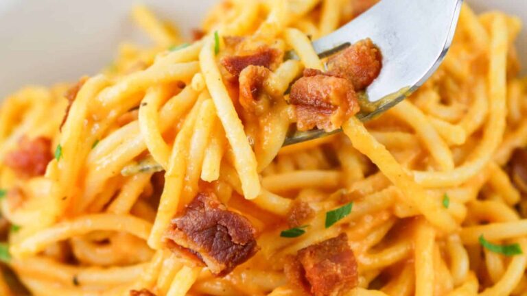 A close-up of spaghetti with a creamy orange sauce, topped with crispy bacon bits and garnished with chopped herbs, with a fork holding some pasta.