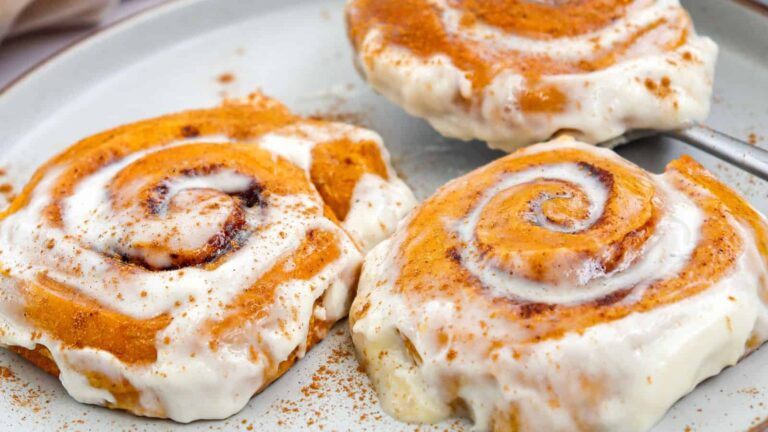 Three cinnamon rolls topped with white icing and a sprinkle of cinnamon are arranged on a plate, with one being served using a spatula.
