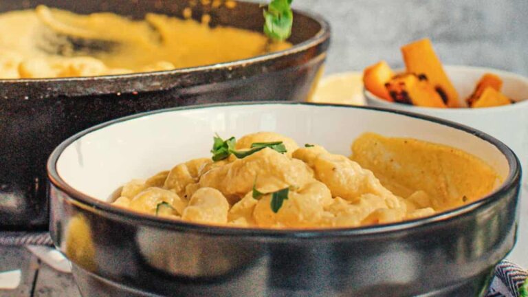 A bowl of creamy pasta garnished with herbs, with a skillet of pasta and a cup of fries in the background.