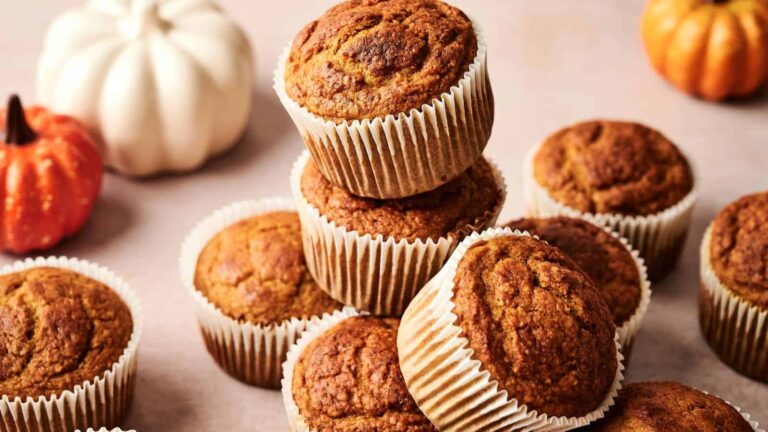 A group of pumpkin muffins in paper liners stacked on a light surface, with small decorative pumpkins in the background.