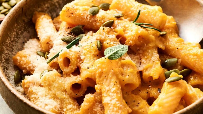 A bowl of rigatoni pasta coated in a creamy orange sauce, topped with grated cheese, pumpkin seeds, black pepper, and fresh herbs.