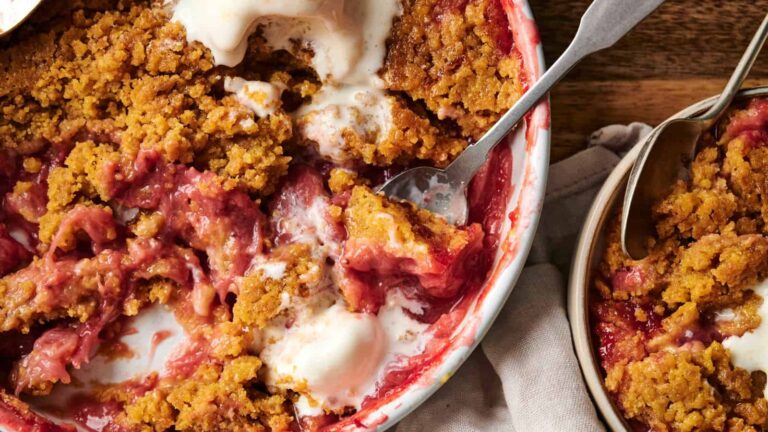 A close-up of a partially served fruit crumble dessert topped with melting vanilla ice cream in a baking dish, with a spoon and a bowl beside it.