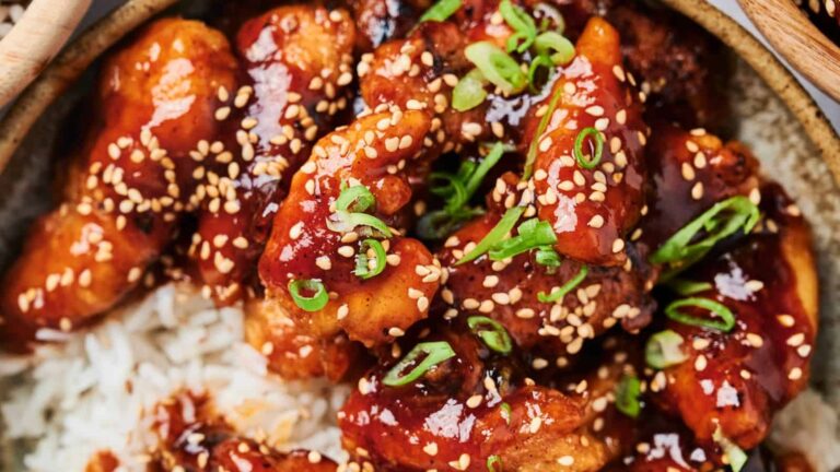 A close-up of glazed chicken pieces topped with sesame seeds and chopped green onions, served over white rice.