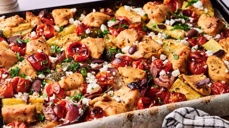 Baked sheet pan dish with chicken pieces, cherry tomatoes, zucchini, olives, and feta cheese, garnished with herbs.