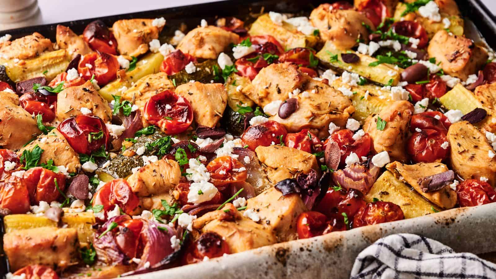 Sheet pan filled with baked chicken pieces, cherry tomatoes, zucchini, olives, and crumbled feta cheese, garnished with herbs.