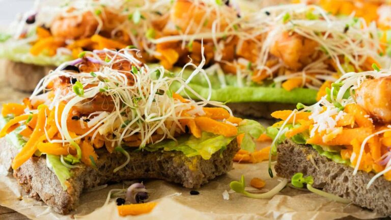 Open-faced sandwiches on whole grain bread topped with lettuce, grated carrots, microgreens, and pieces of cooked shrimp, arranged on parchment paper.