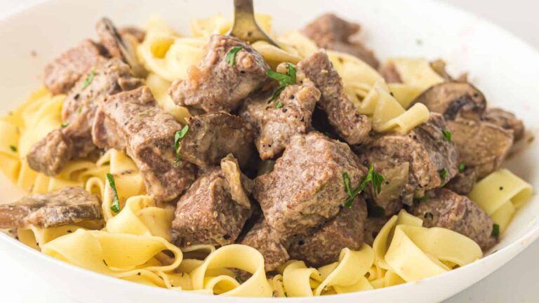A plate of egg noodles topped with chunks of beef in a creamy sauce, garnished with herbs, with a fork twisting some pasta.