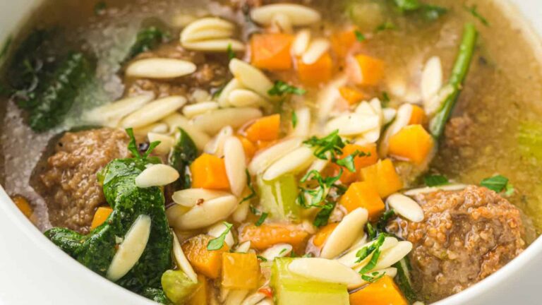A close-up of a bowl of soup with meatballs, orzo pasta, chopped carrots, celery, spinach, and fresh herbs in a clear broth.
