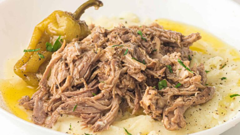 Shredded beef atop mashed potatoes, garnished with chopped herbs and a whole pepper, served with a light broth.