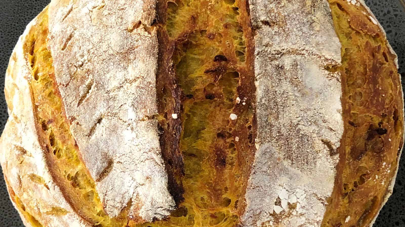 A close-up of a round, crusty homemade loaf of bread with a golden-brown color and two deep slashes across the top, dusted lightly with flour.
