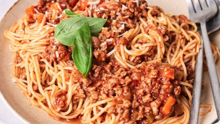 A plate of spaghetti topped with meat sauce, garnished with fresh basil leaves, with a fork and spoon placed on the side.