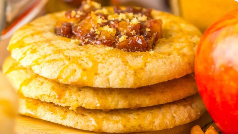 Three stacked cookies with a crumbly texture, each topped with a chunky fruit filling, next to a red apple.