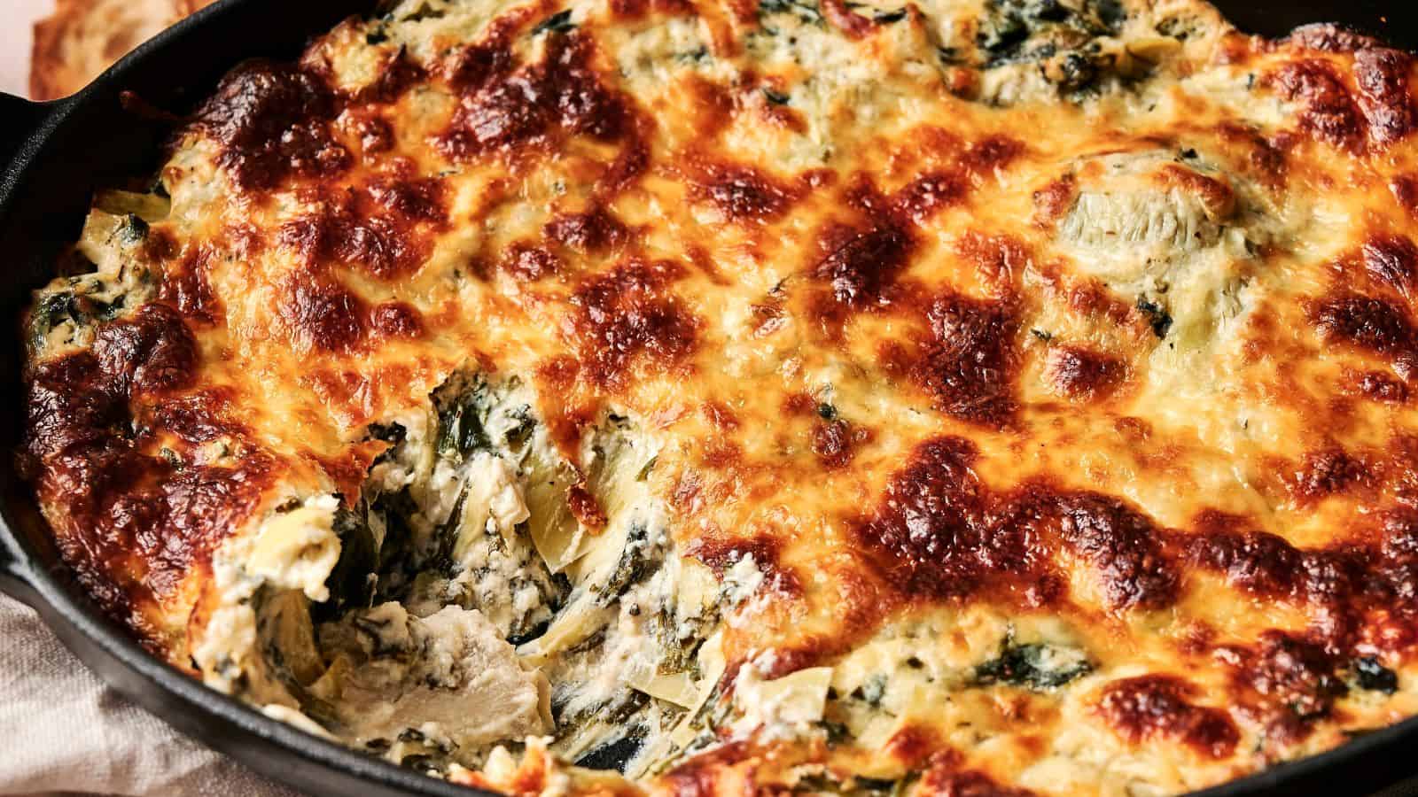 Close-up of a baked spinach and artichoke dip in a dark dish, showing a golden brown, bubbly cheese topping with a portion scooped out, revealing the creamy interior.