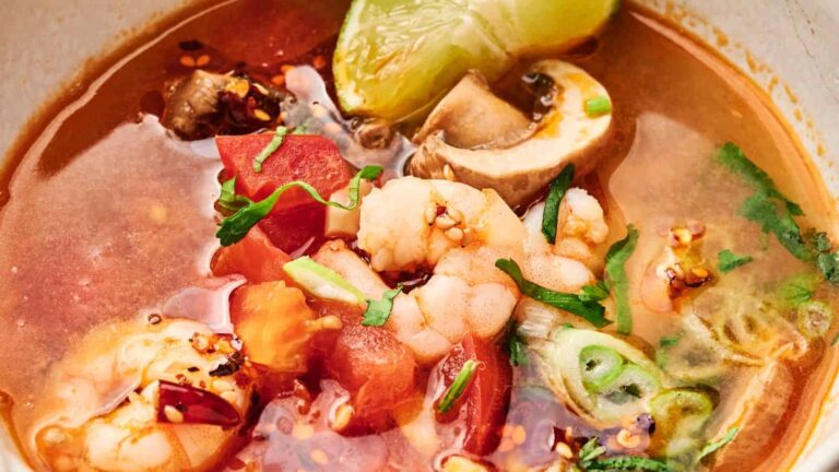 A bowl of spicy seafood soup with shrimp, mushrooms, tomatoes, green onions, fresh herbs, and a lime wedge in a red broth.
