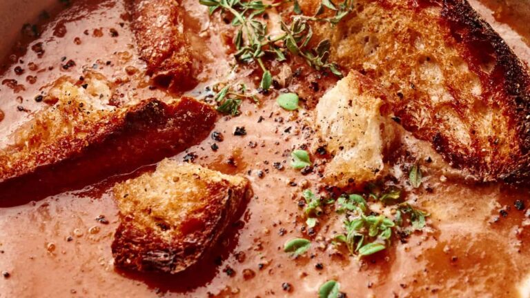 Close-up of tomato soup topped with crispy toasted bread pieces, black pepper, and fresh thyme leaves.