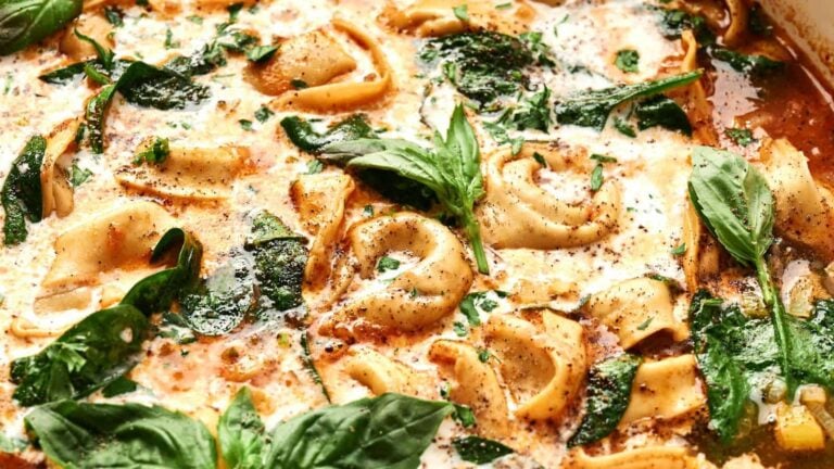 Close-up of creamy tortellini pasta with spinach and fresh basil leaves, topped with black pepper and herbs.