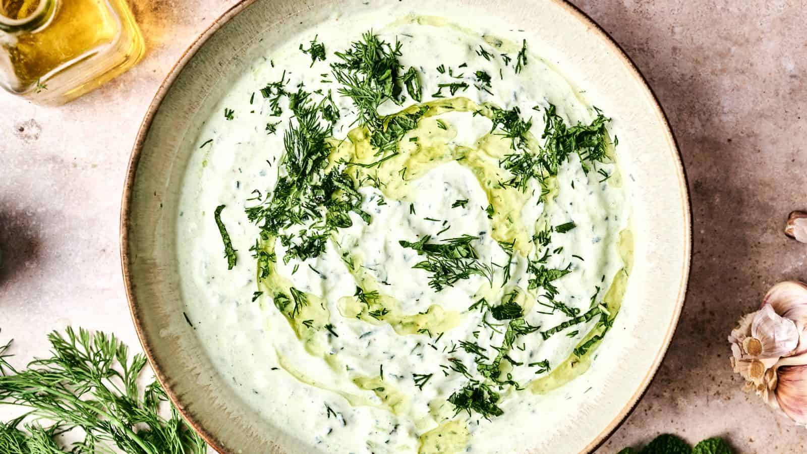 A bowl of creamy tzatziki sauce garnished with chopped dill and a swirl of olive oil. Fresh dill and garlic are nearby.