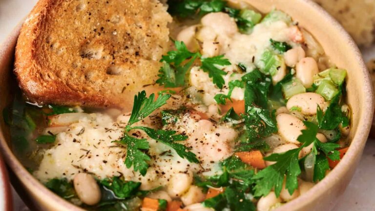A bowl of white bean and vegetable soup topped with melted cheese, fresh parsley, and a slice of toasted bread.