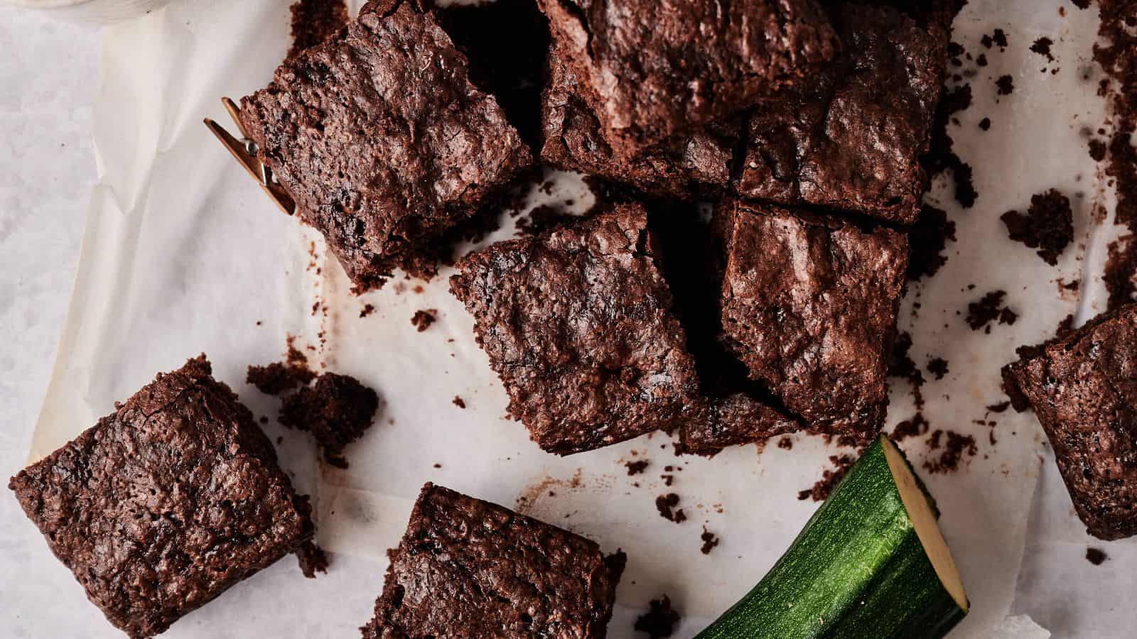 Several brownies are arranged on parchment paper with crumbs scattered around and a piece of zucchini placed next to them.