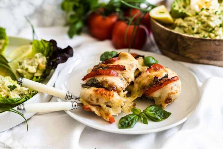 A white plate with baked chicken breasts stuffed with cheese, tomatoes, and herbs, garnished with basil, beside a salad and a bowl of rice.