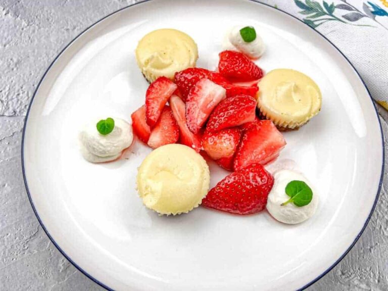 A plate with three mini cheesecakes, fresh sliced strawberries, and four dollops of whipped cream topped with small green leaves.
