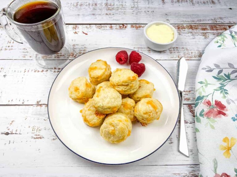 A plate of Mini Flaky Cheese Biscuits with raspberries, accompanied by a glass mug of coffee and a small bowl of yellow sauce on a white wooden table with a floral napkin.