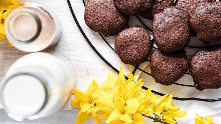Chocolate cookies on a cooling rack next to two bottles of milk and yellow flowers on a white surface.