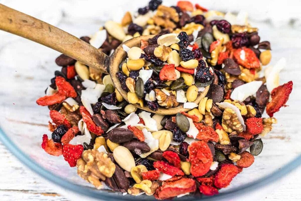 A wooden spoon scoops a mix of dried fruits, nuts, seeds, and chocolate chips from a glass bowl on a white surface.