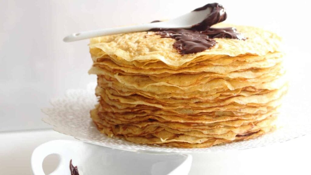 A stack of thin crepes on a plate with a white spoon resting on top, covered in a dollop of chocolate spread.
